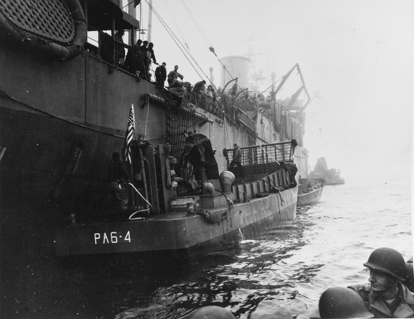 Black and white photograph of soldiers in helmets in a landing craft alongside a large transport ship in fog, with an American flag at the stern.