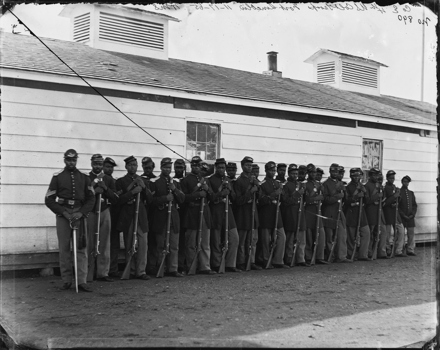 Black-and-white photograph of a line of Black soldiers in Union uniform with rifles, a sergeant with a sword at the left, in front of a white wooden barracks.