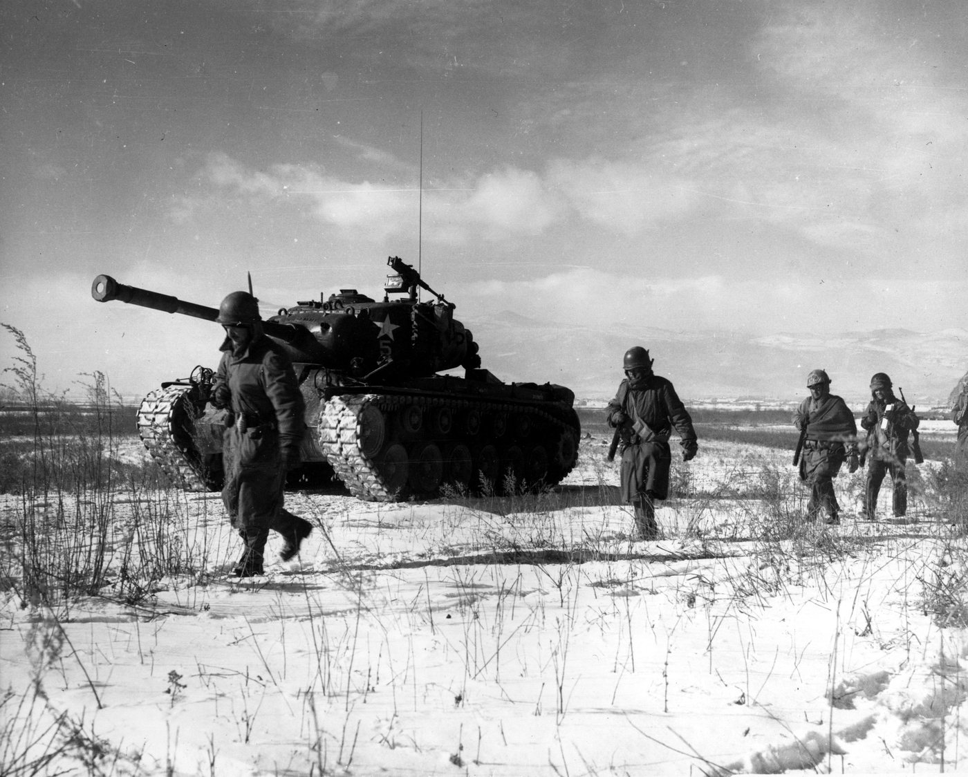 Black and white photograph of Marines walking beside a tank across a snowy field in Korea