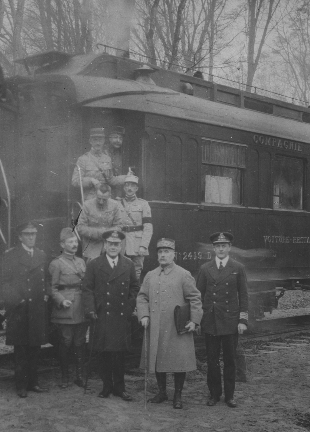 Officers in greatcoats and naval uniforms stand in front of a dark railway dining car marked Compagnie in a bare winter forest, with more officers on the carriage steps behind them