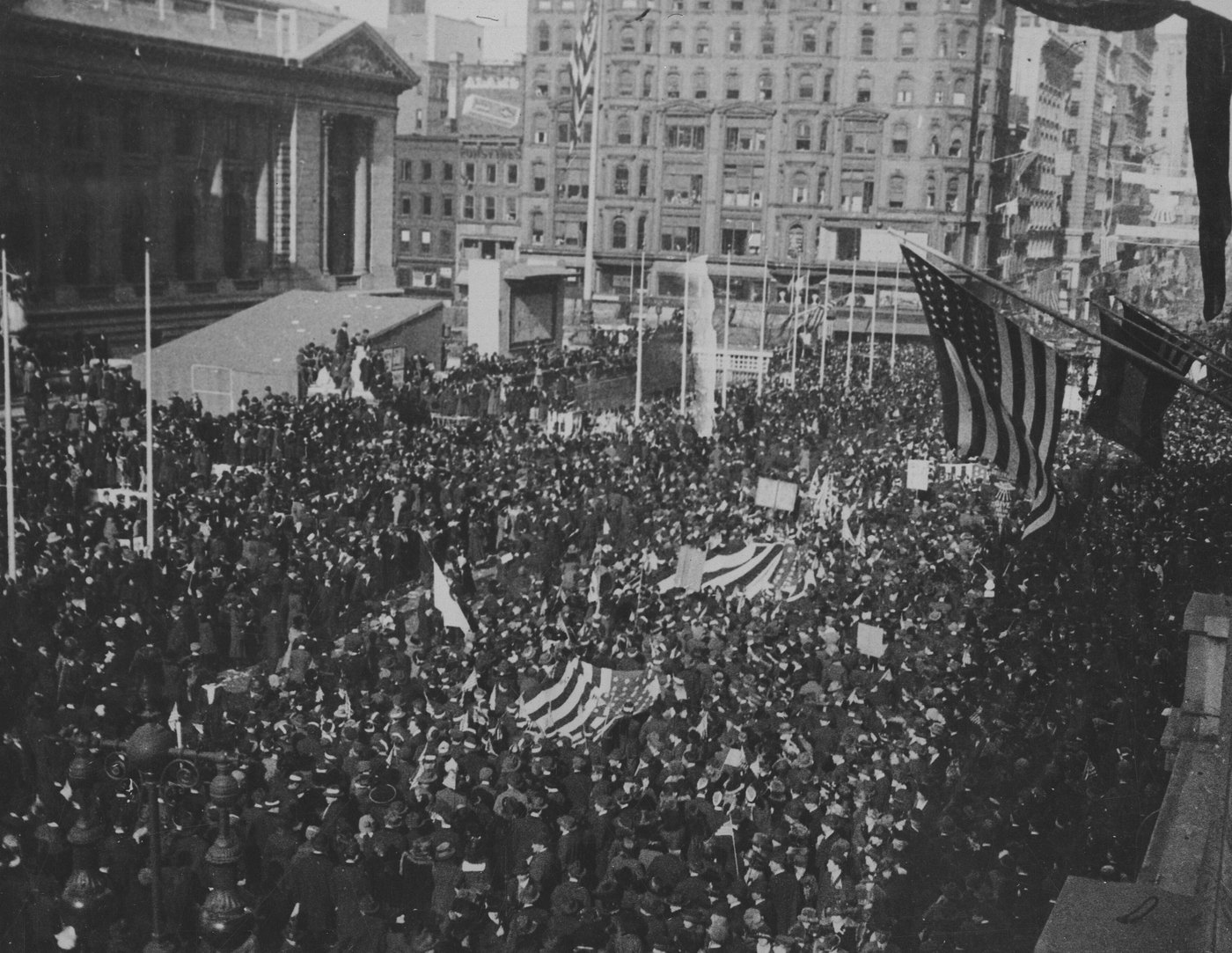 A dense crowd fills a city street in front of a large classical library building, seen from above, with American flags hanging from a building and carried among the people