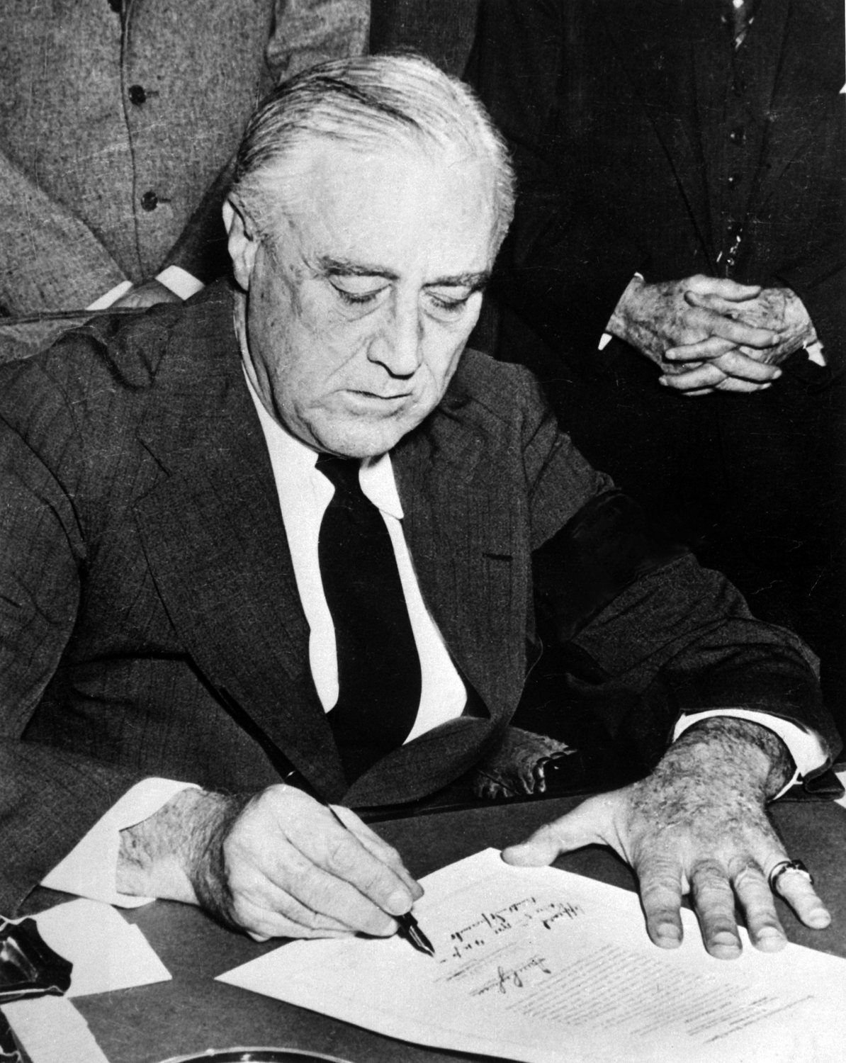Black and white photograph of Franklin Roosevelt seated at a desk signing a document with a fountain pen