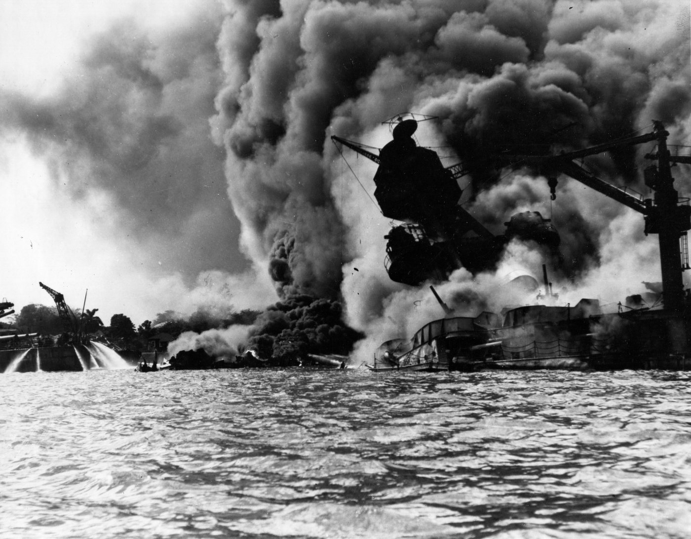 Black and white photograph of a battleship engulfed in billowing black smoke, its tripod mast leaning