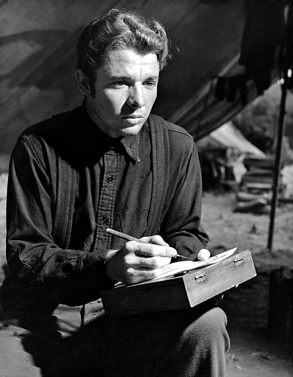 Black and white photograph of a young Audie Murphy seated and writing on a wooden box