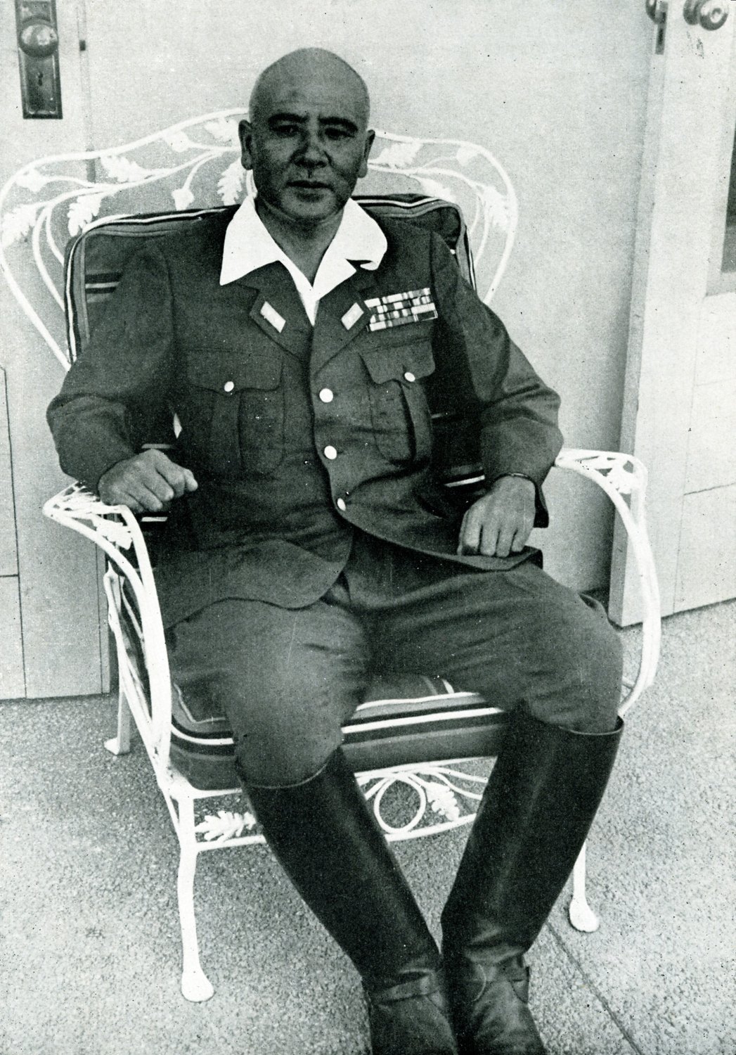 Black and white photograph of a shaven headed Japanese general in a field uniform with medal ribbons and tall riding boots, sitting in a white wrought iron garden chair on a porch