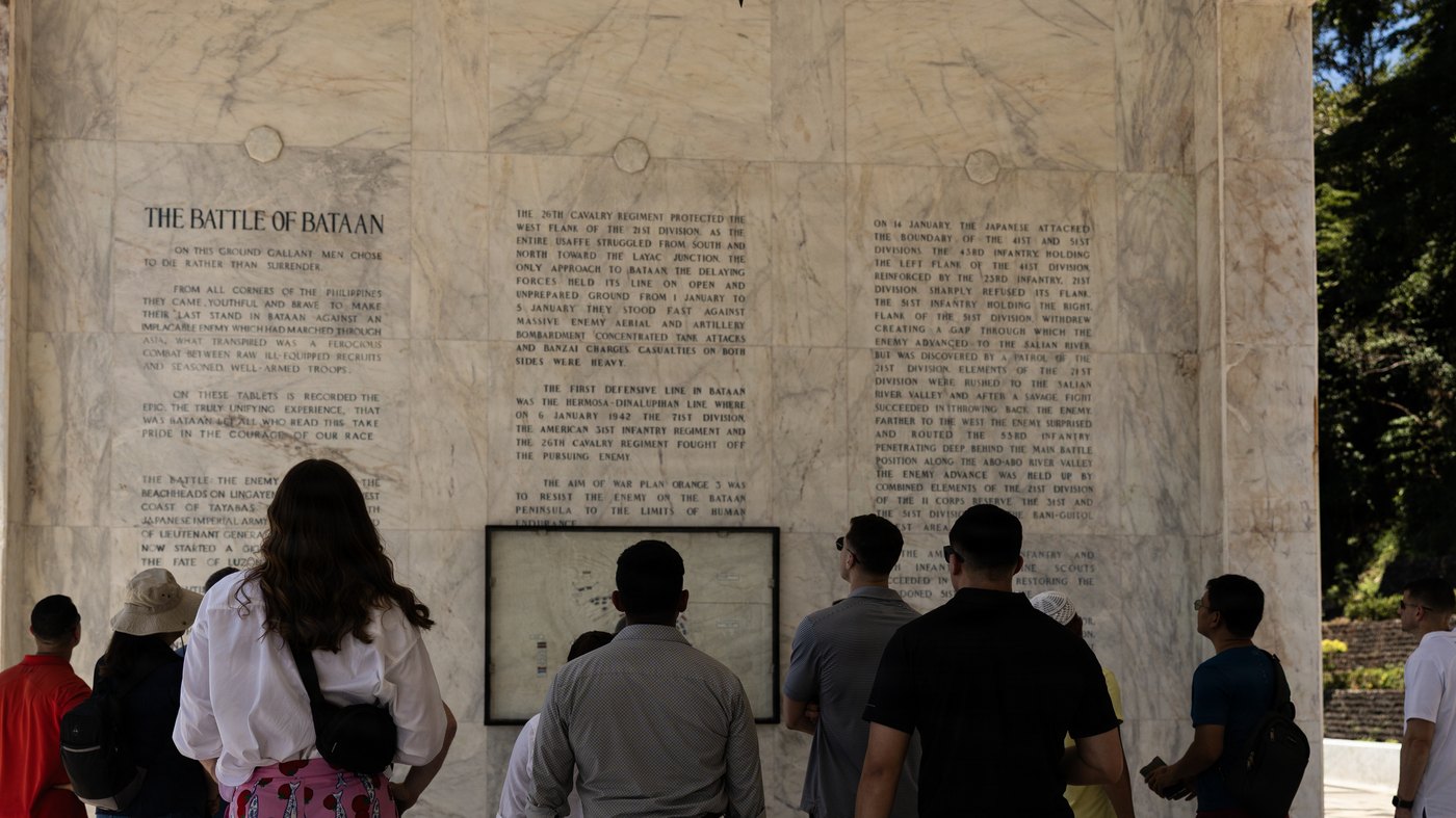 Visitors stand before a wall of white marble tablets engraved with columns of text under the heading The Battle of Bataan, with a framed map between the panels