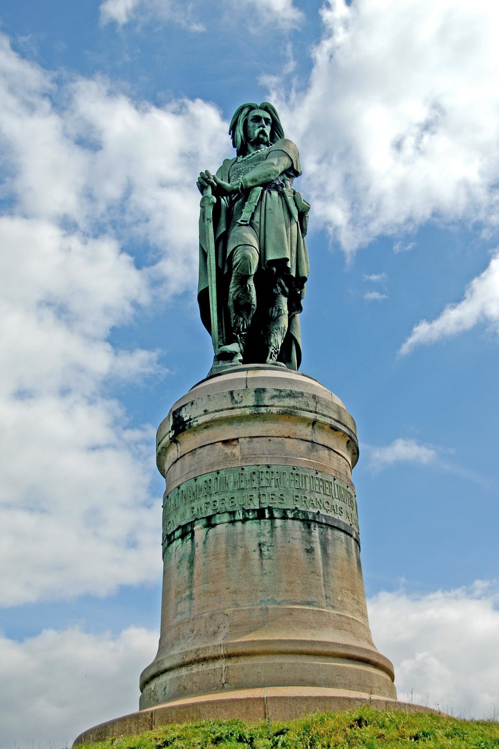 Tall hammered copper statue of a Gallic warrior on a high stone pedestal against a blue sky