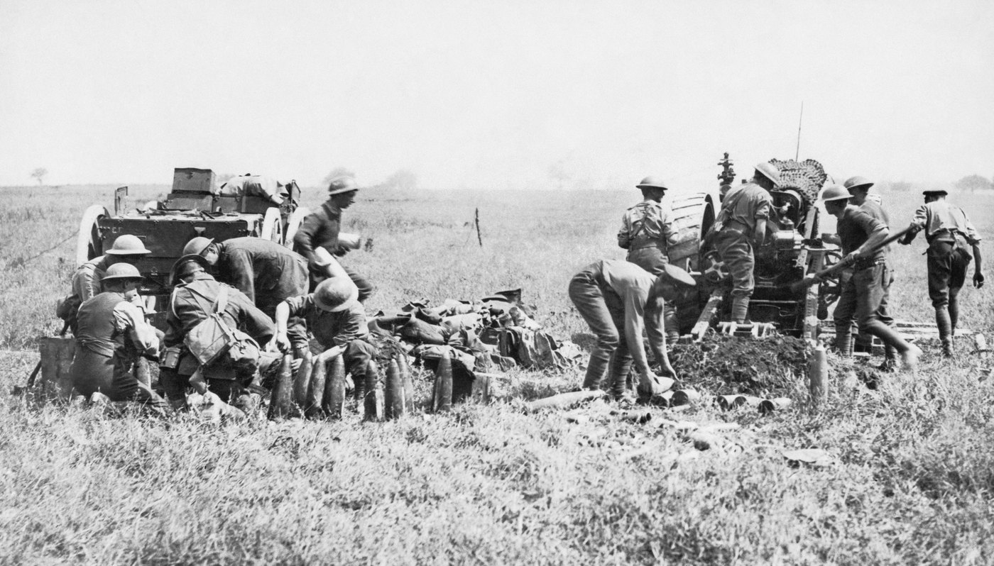 Black and white photograph of British heavy field guns firing in an open grassy field, gunners in steel helmets loading one gun while others crouch beside a row of large shells and an ammunition wagon on the left