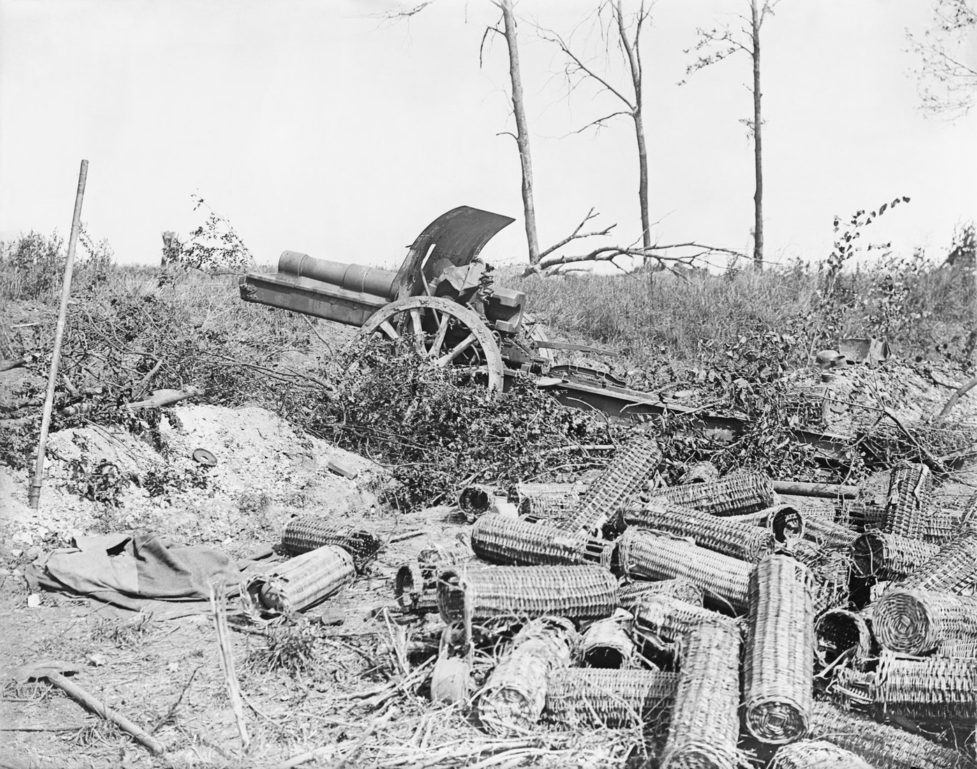 Black and white photograph of an abandoned German field howitzer among broken branches on a slope, with a large heap of empty wicker shell carriers in the foreground and bare shattered trees behind