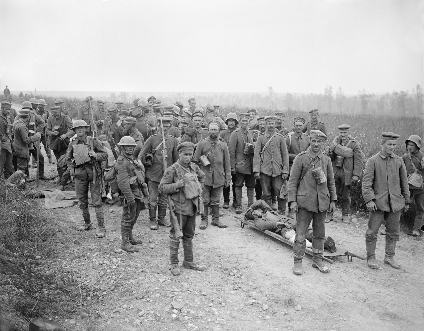 Black and white photograph of a large crowd of German prisoners in field caps and long coats standing on a country road, some holding mess tins, with a wounded man on a stretcher in front and a few British soldiers in steel helmets guarding them
