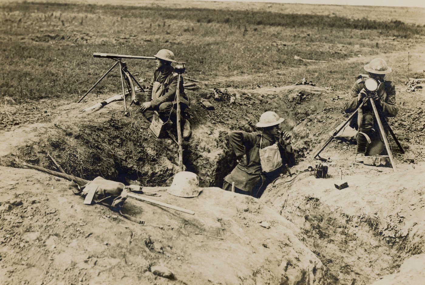 Sepia photograph of three British soldiers in a captured trench on open ground, one seated beside a heliograph on a tripod, one in the trench using a telephone handset and one kneeling behind a signal lamp on a tripod, with a German helmet lying on the parapet