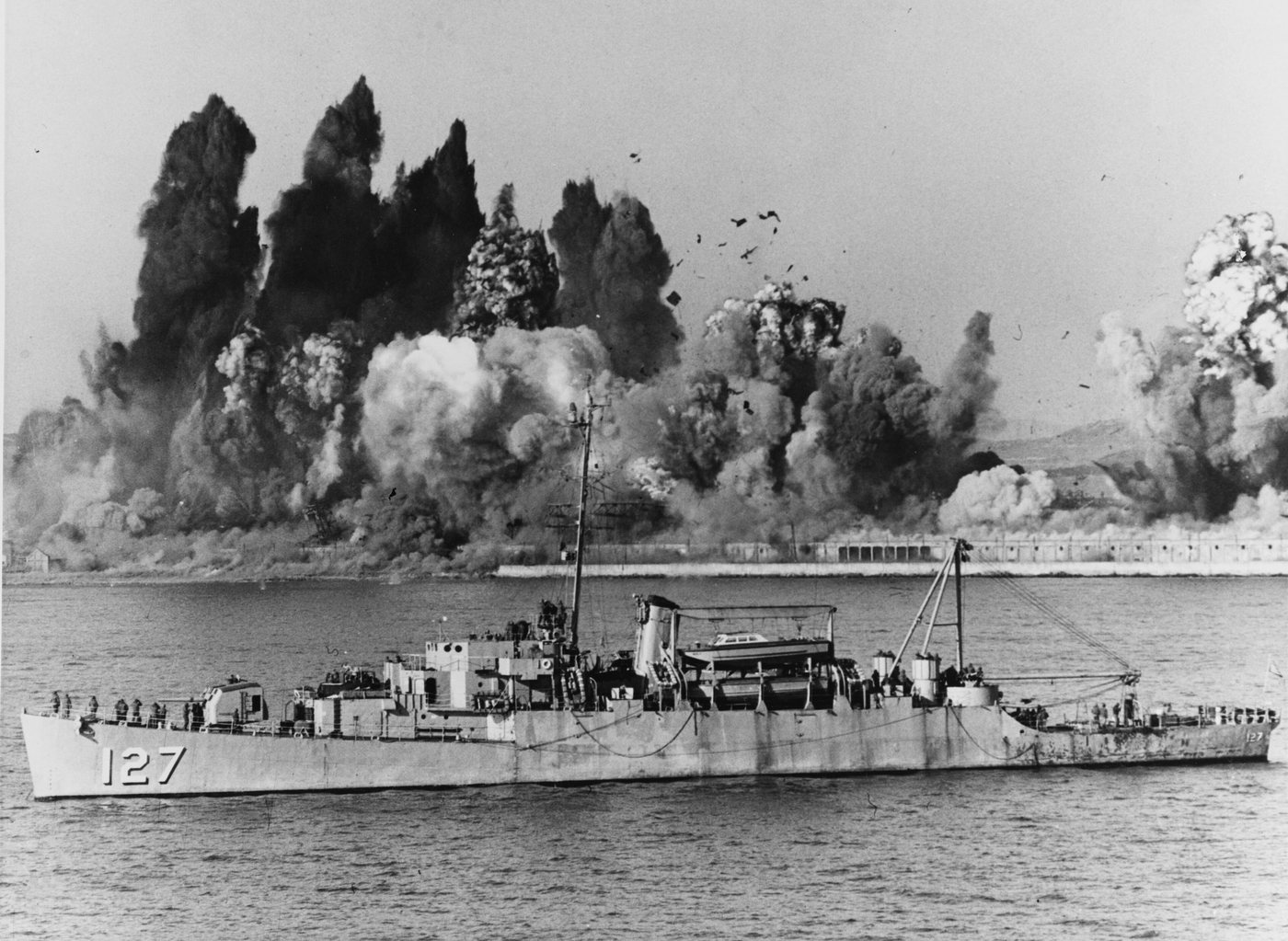 Photograph of a huge explosion cloud over a harbour with a warship in the foreground