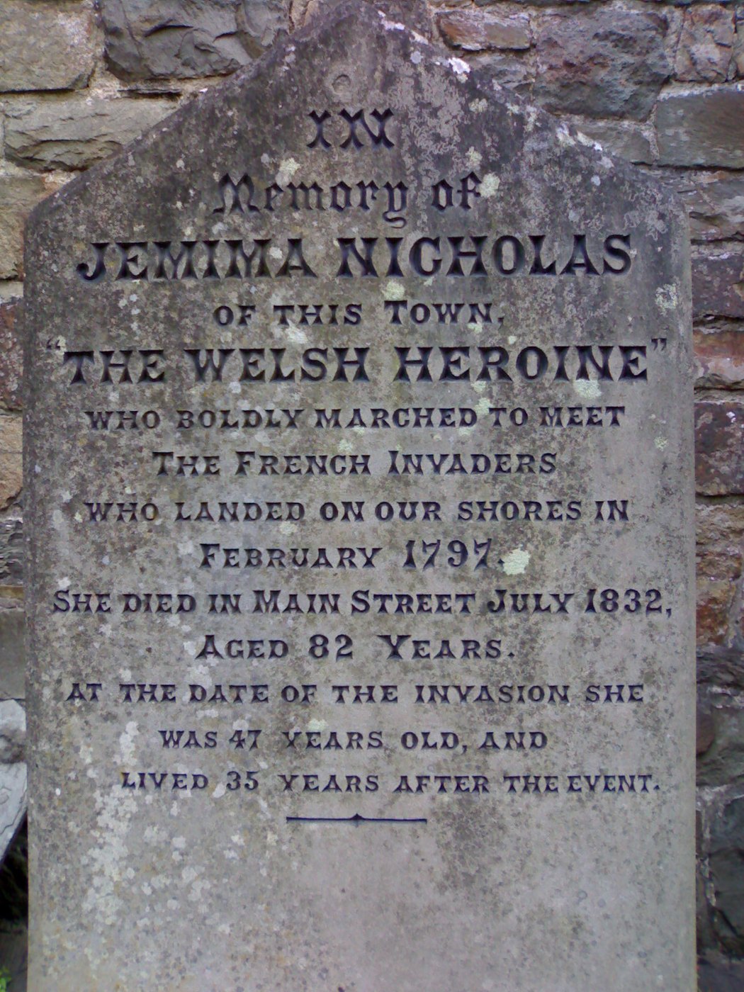 Zwietrzały szary kamień pamiątkowy przy kamiennym murze z inskrypcją In memory of Jemima Nicholas of this town, the Welsh heroine who boldly marched to meet the French invaders who landed on our shores in February 1797