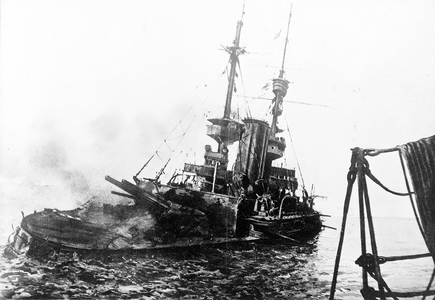 Photograph of a listing battleship low in the water, abandoned by its crew