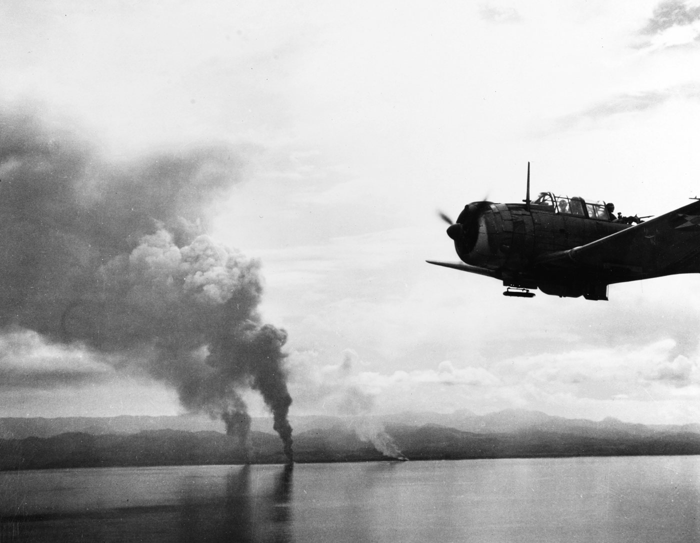 Aerial photograph of burning ships run aground on a beach with a dive bomber in the foreground
