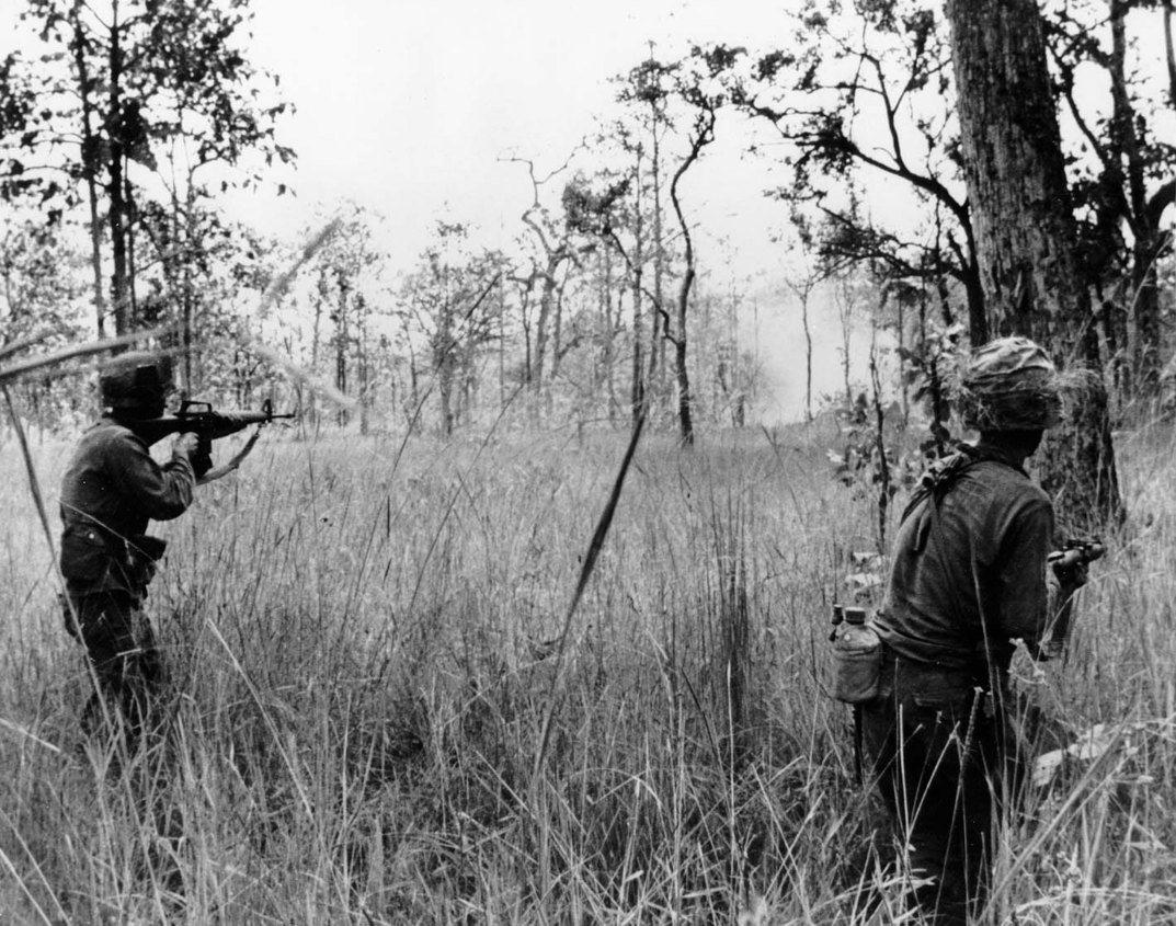 Photograph of two American soldiers with rifles in tall grass among scattered trees