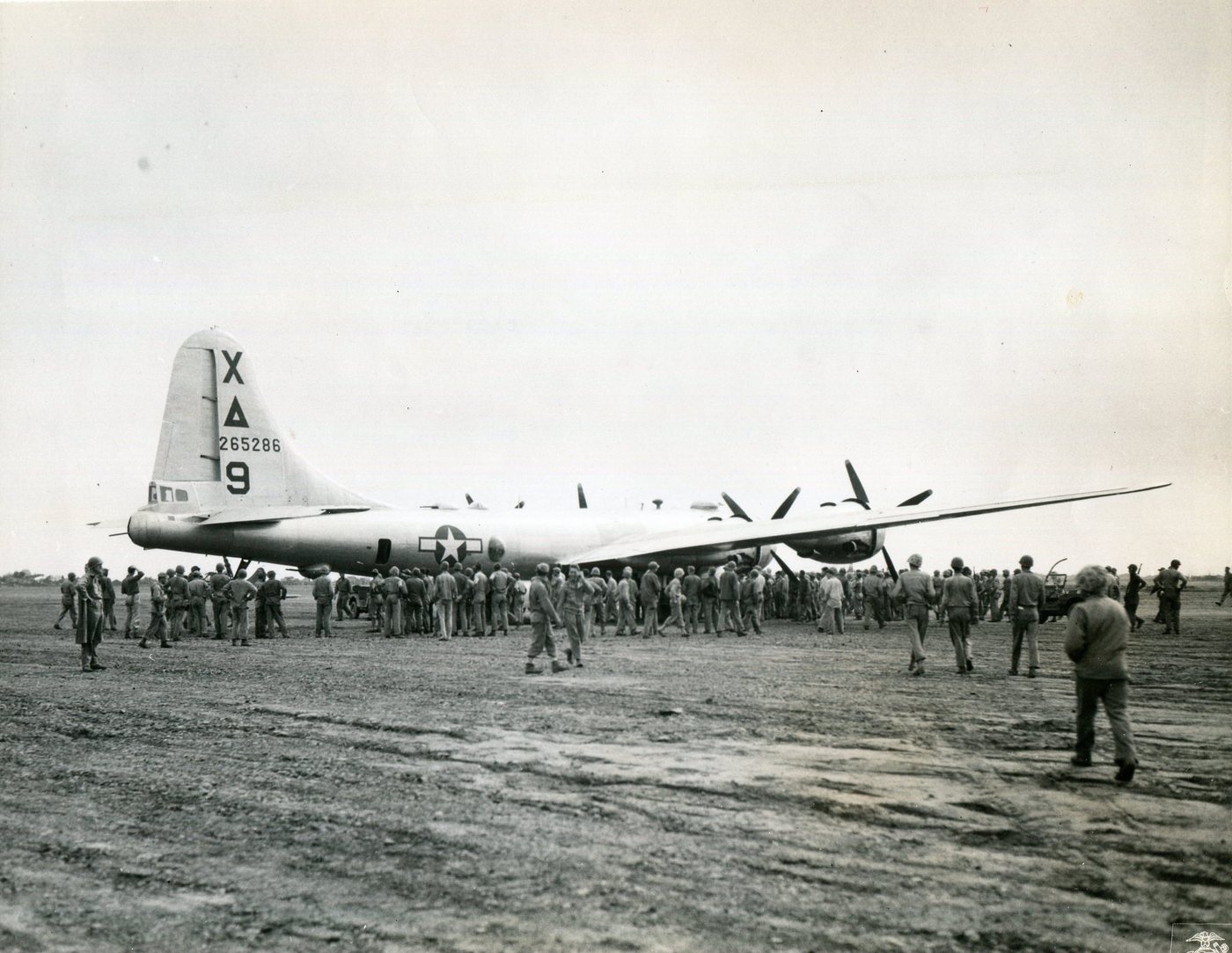Large four-engined bomber parked on a dirt airfield with a crowd of servicemen around it