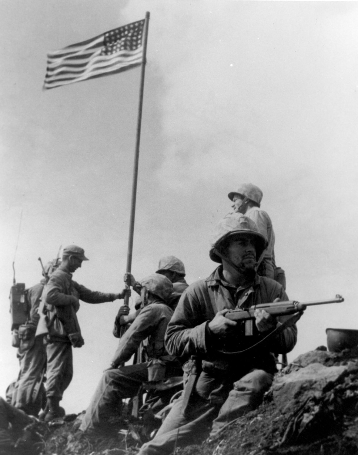 Marines on a rocky summit holding up a small American flag on a pole while one keeps watch with a carbine