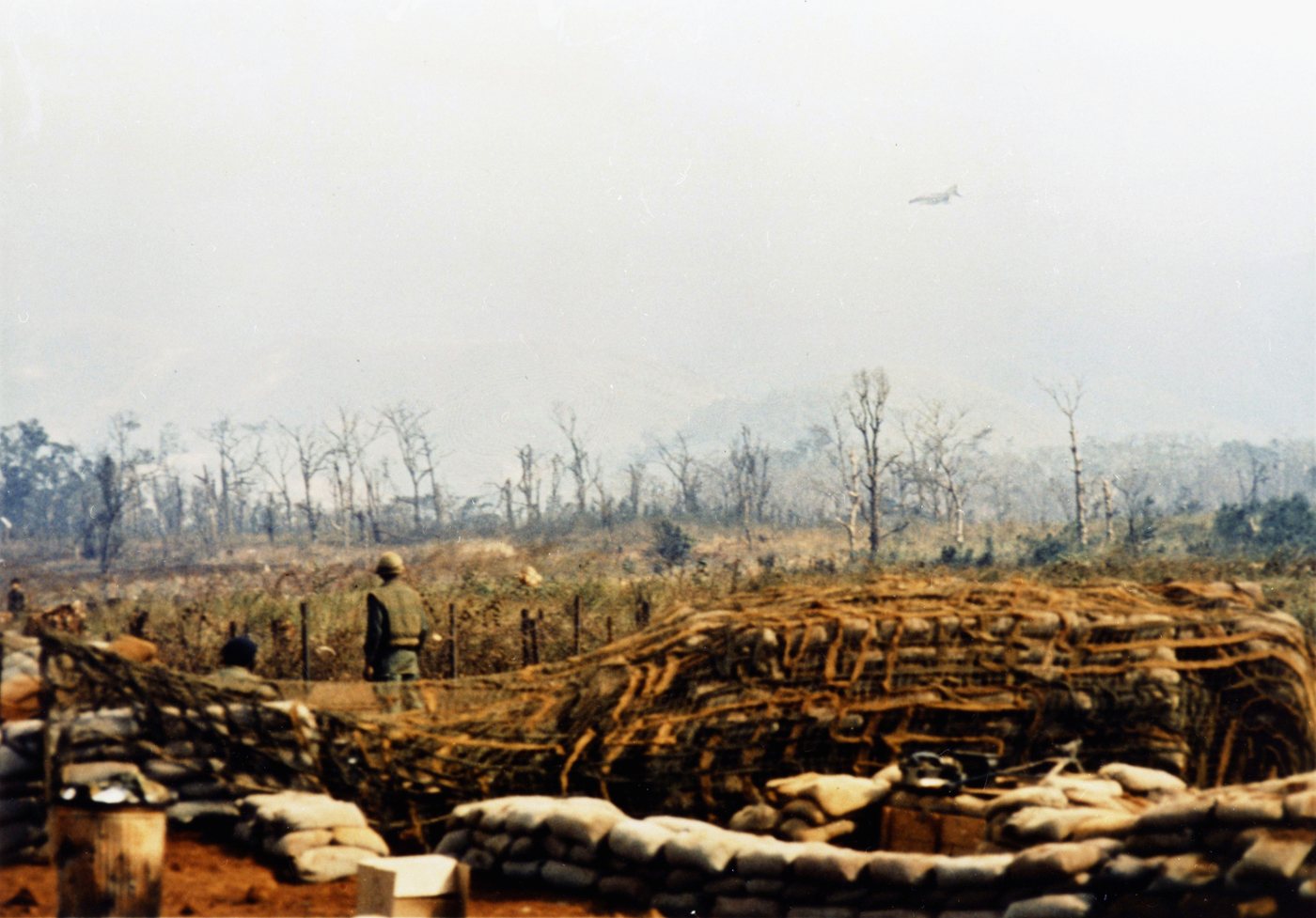 Sandbagged bunkers in a scarred landscape with a jet aircraft flying low in the distance