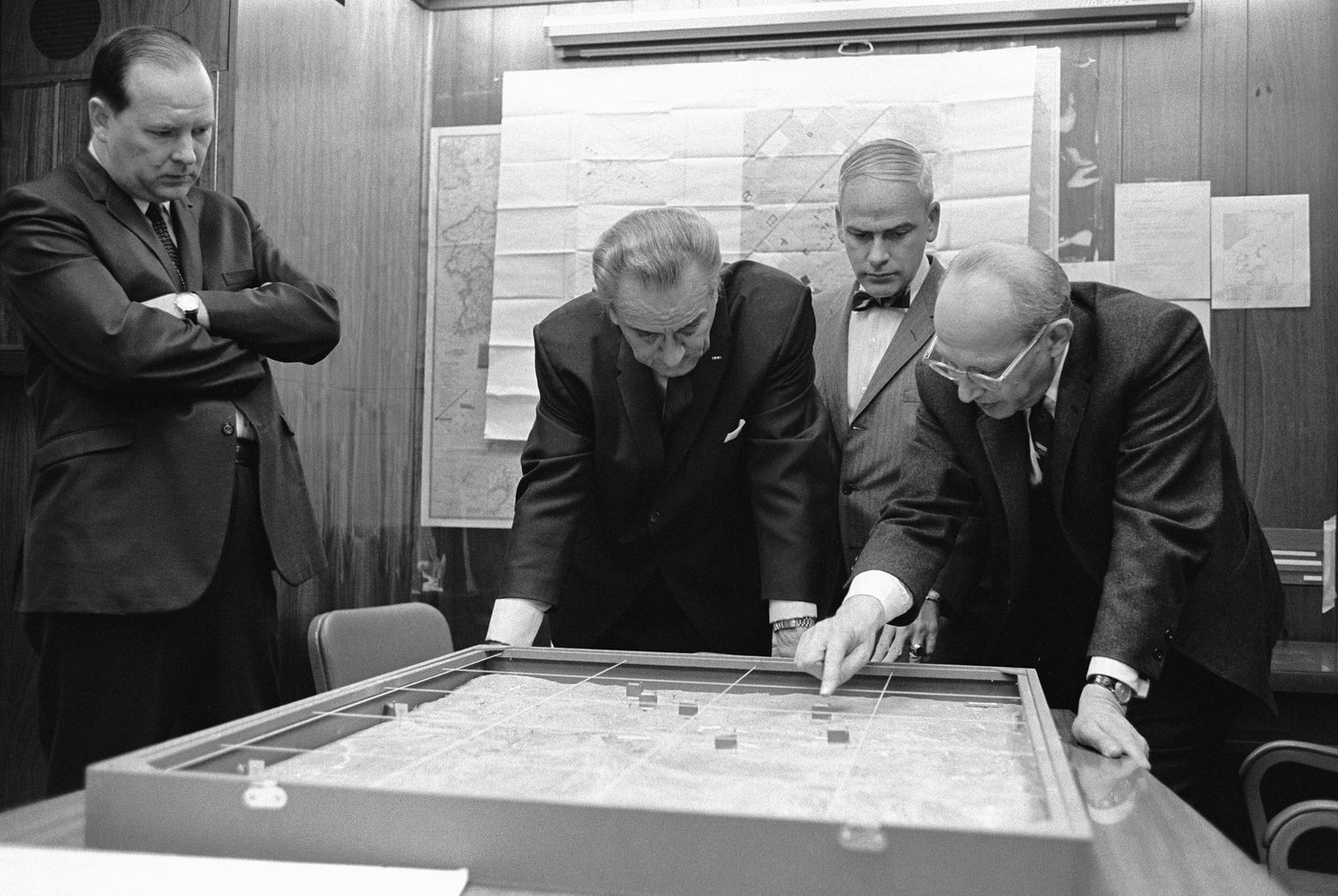 Men in suits bending over a terrain model on a table in front of wall maps