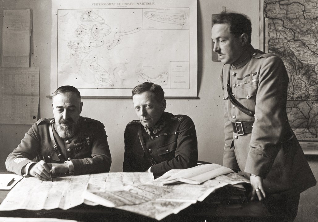 Trzech oficerów w mundurach przy stole zasłanym mapami, jeden stoi