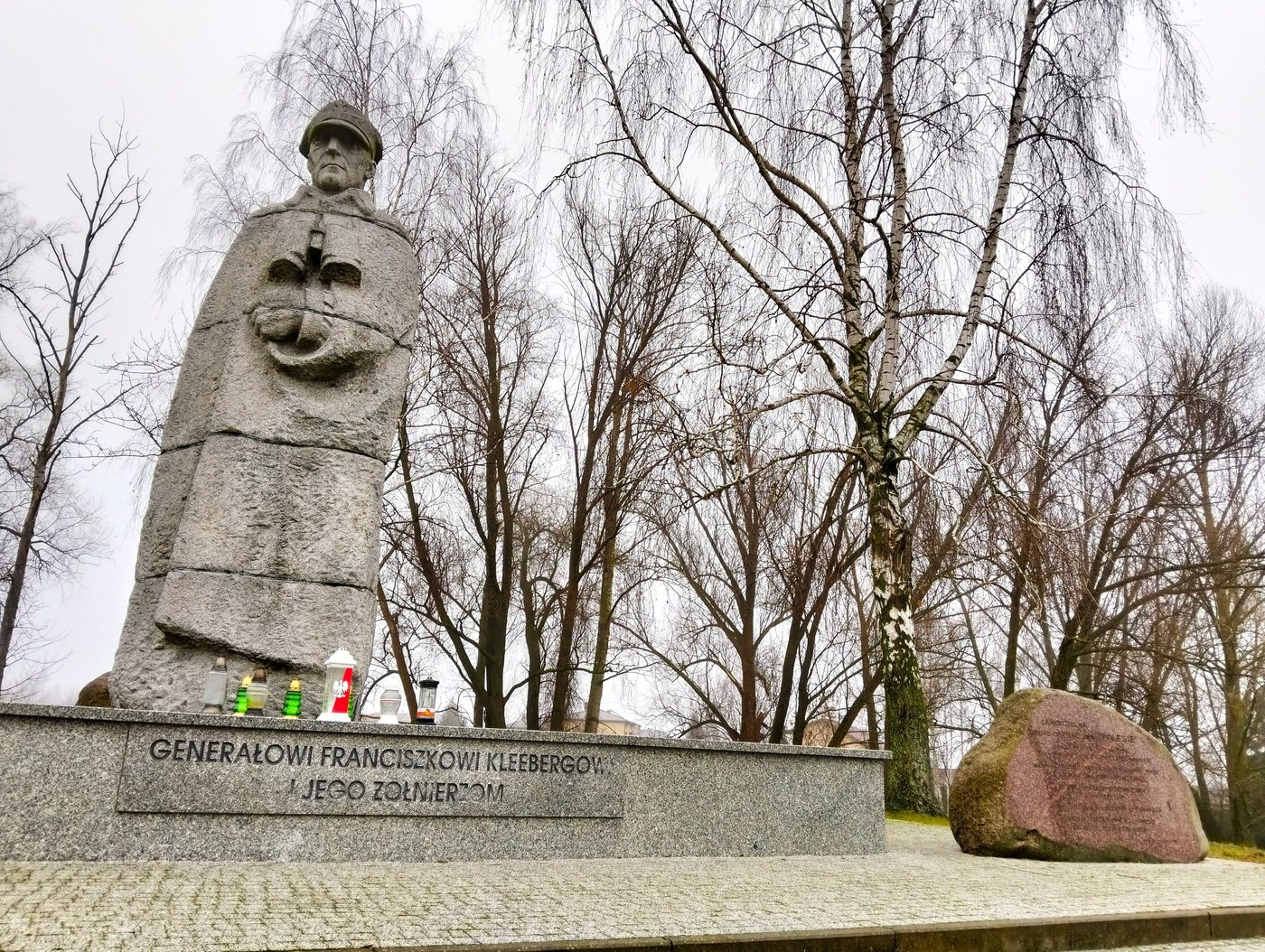 Kamienny posąg generała w płaszczu na granitowym cokole z kwiatami i zniczami