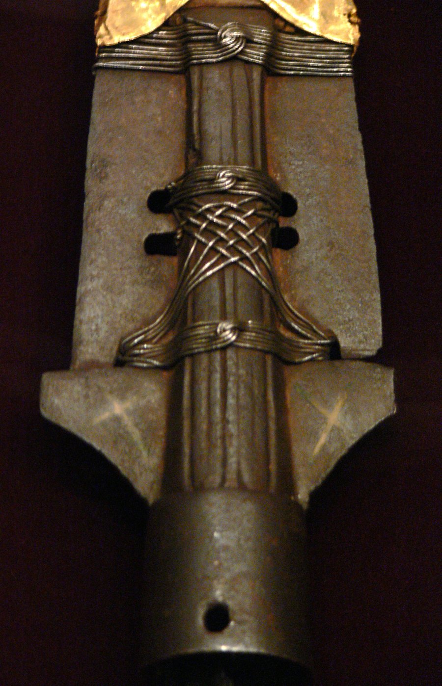Close-up photograph of an old iron spearhead on a dark background, with silver wire wound around its center and a gold sleeve at the top