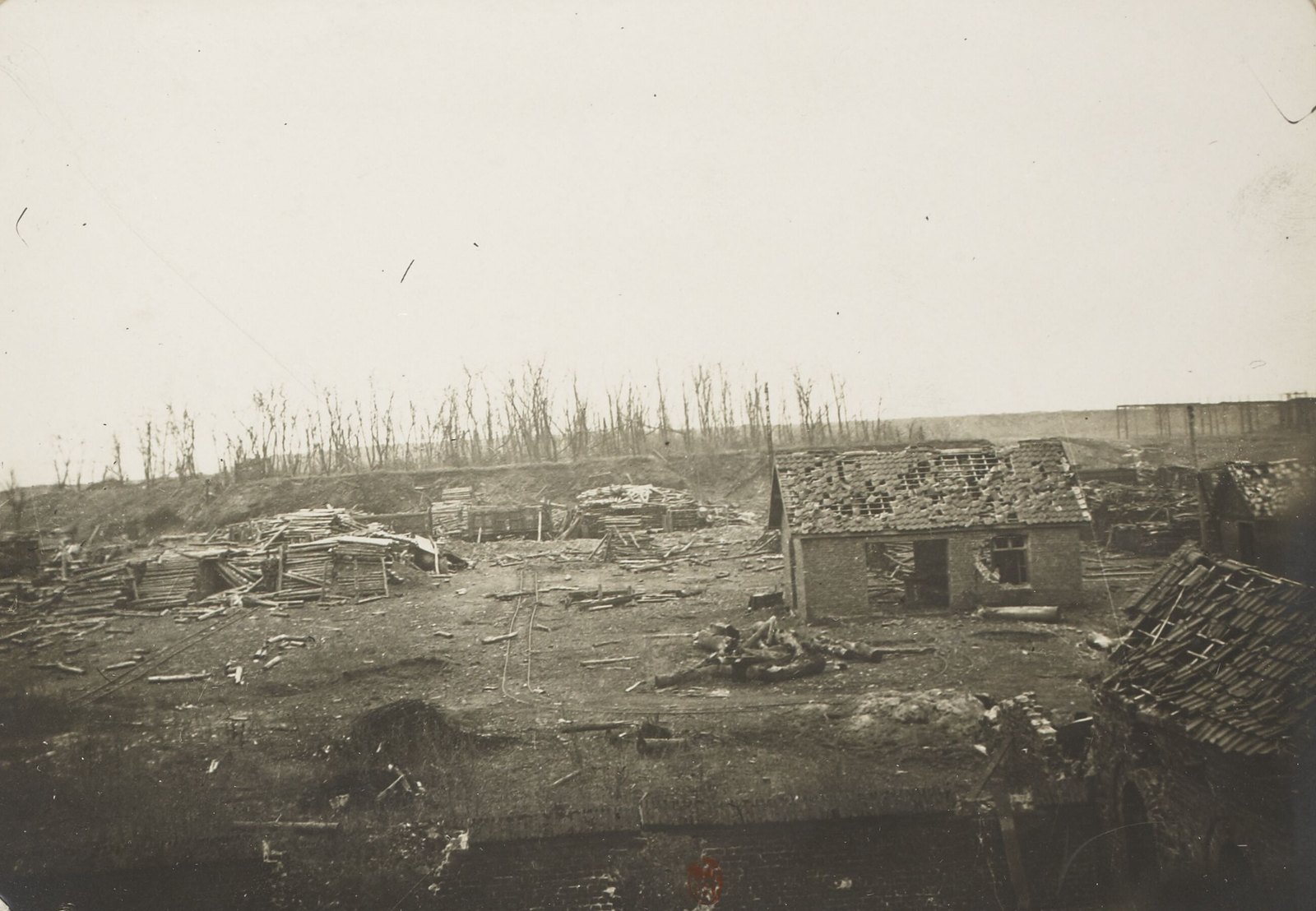 Sepia photograph of a wrecked mining yard littered with timber and debris, a roofless brick hut in the middle and shattered tree stumps along an embankment behind