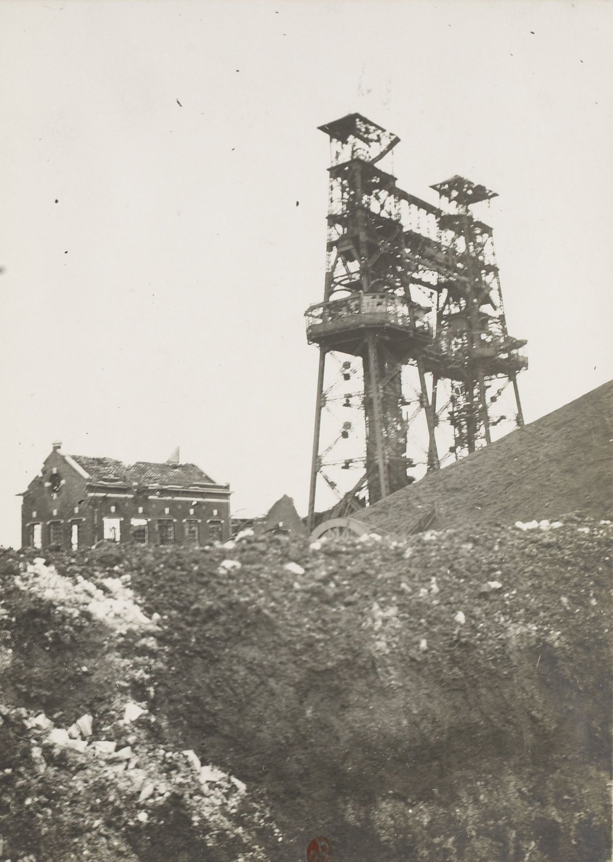 Sepia photograph of a tall double steel pithead headframe beside a slag heap, with a damaged brick mine building at left and churned earth in the foreground