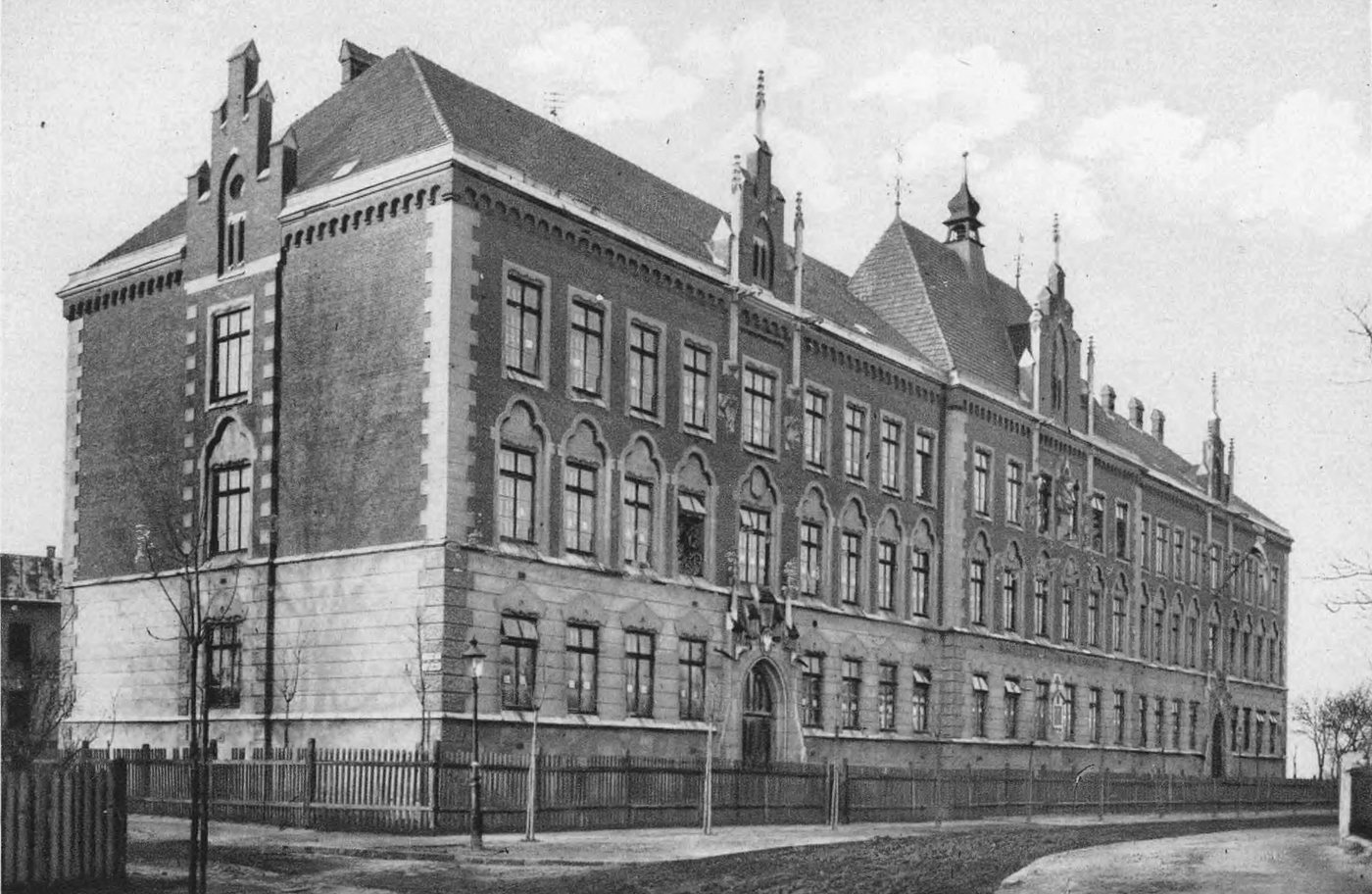 Czarno-białe zdjęcie dużego ceglanego budynku szkoły ze szczytami i ogrodzeniem