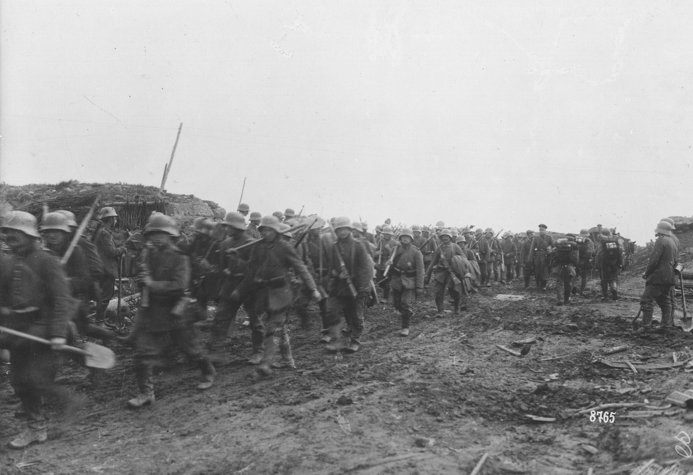 Black and white photograph of a long column of German soldiers in steel helmets, carrying rifles and spades, marching along a muddy track past a ruined earthwork under a gray sky