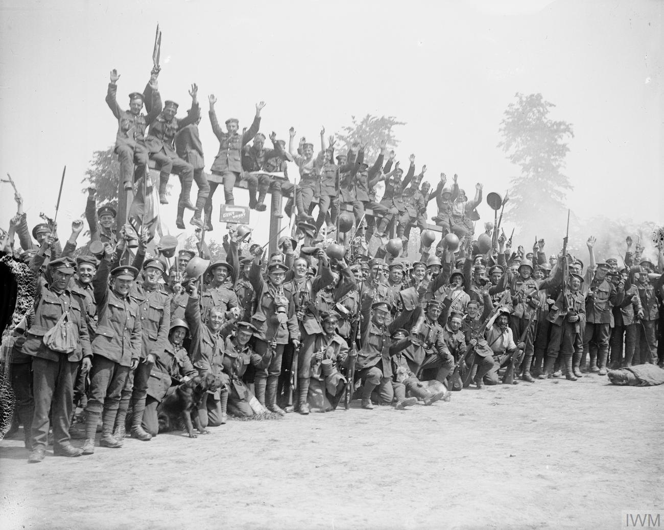 Black and white photograph of a large crowd of cheering British soldiers, many waving arms, rifles and captured German helmets, some sitting on top of a wooden signpost structure, with a dog in the front row