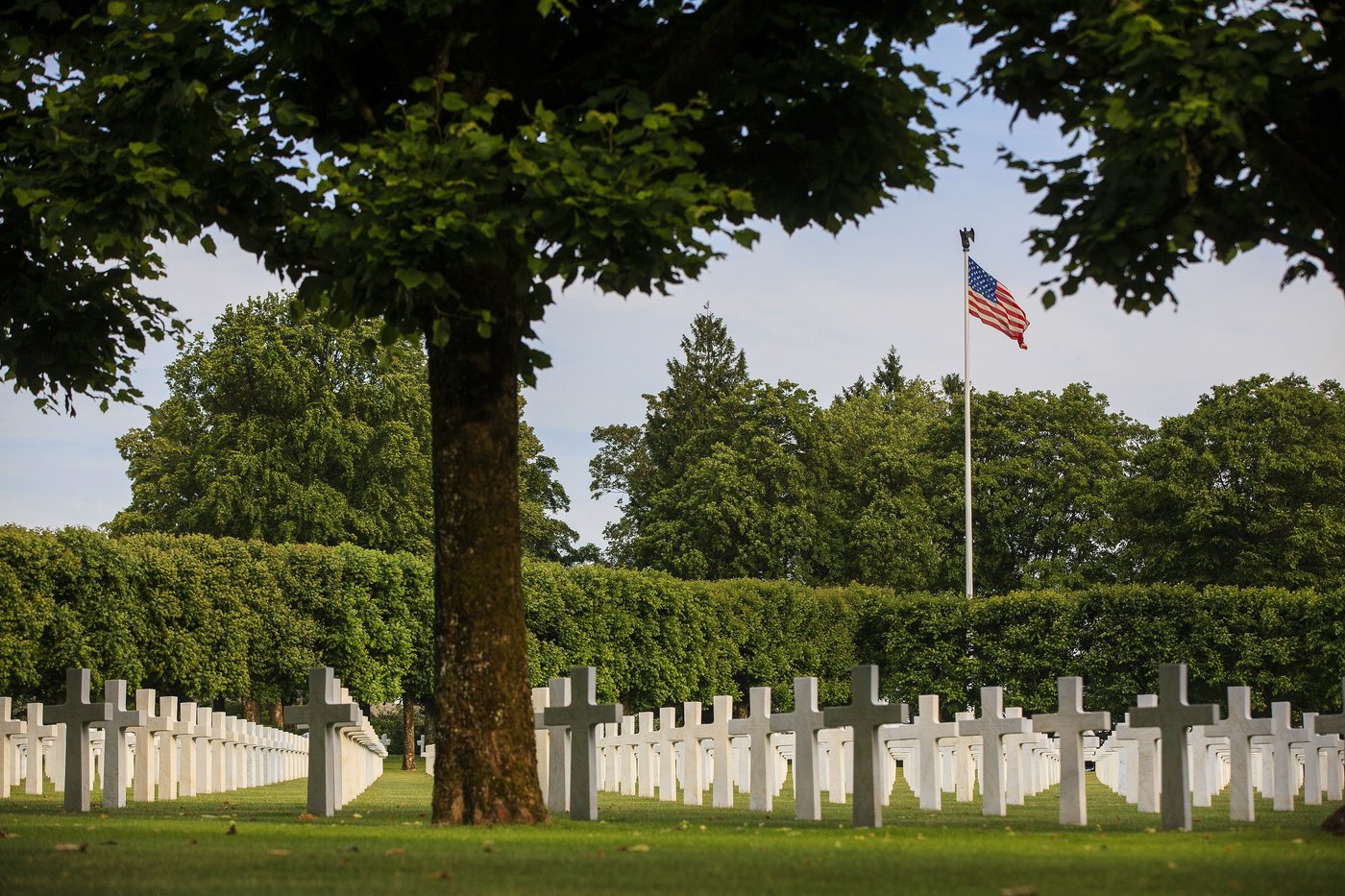 Long rows of white marble crosses on a lawn with an American flag behind the trees