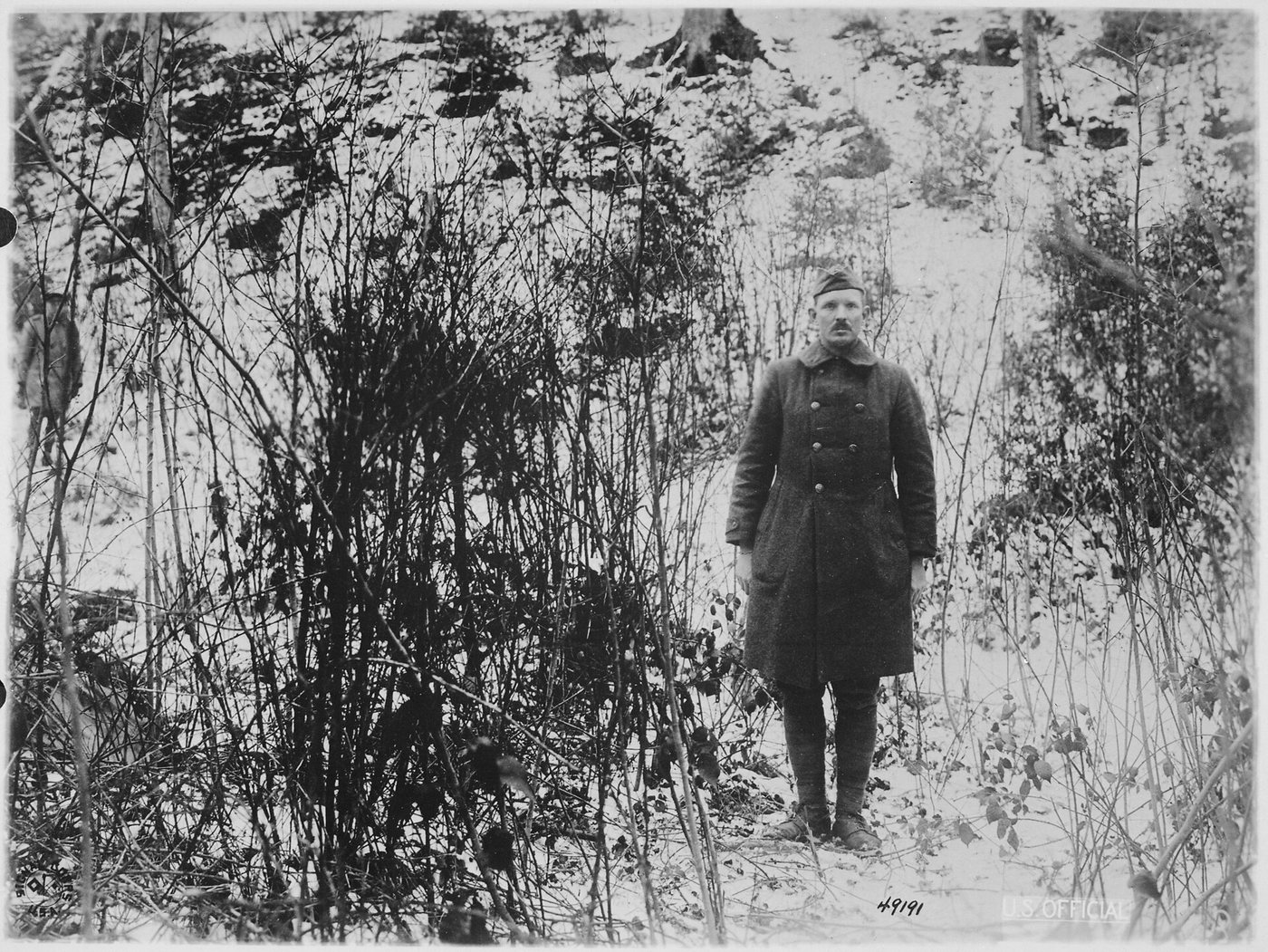 Black and white photograph of a soldier in a greatcoat standing among bare trees on a snowy hillside