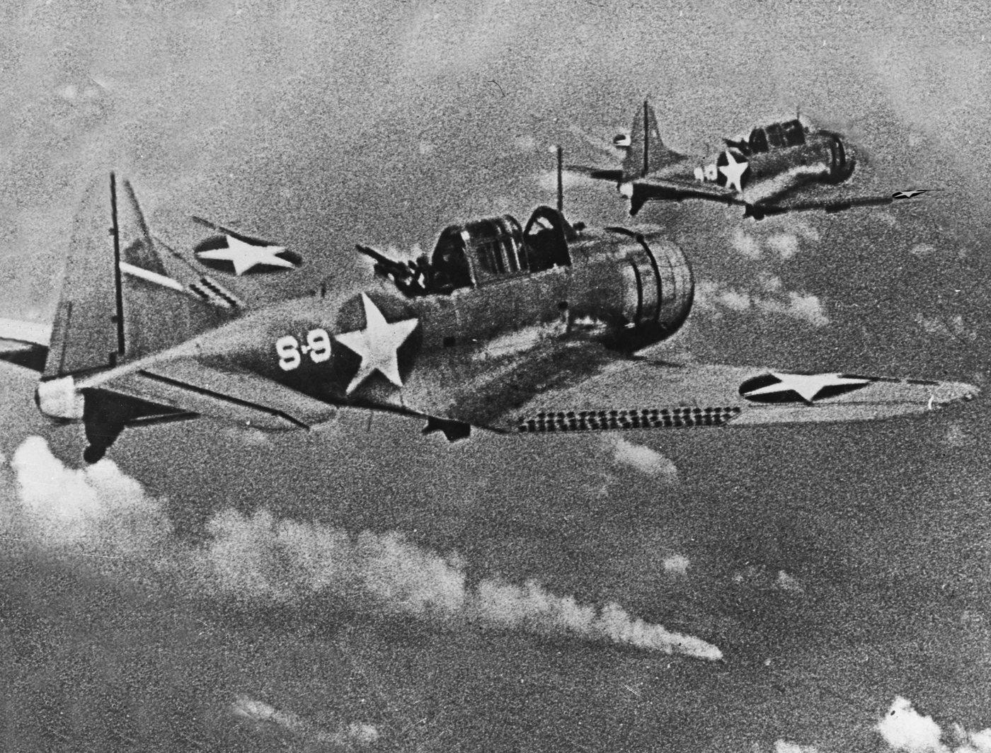 Two US Navy dive bombers in flight above a burning ship