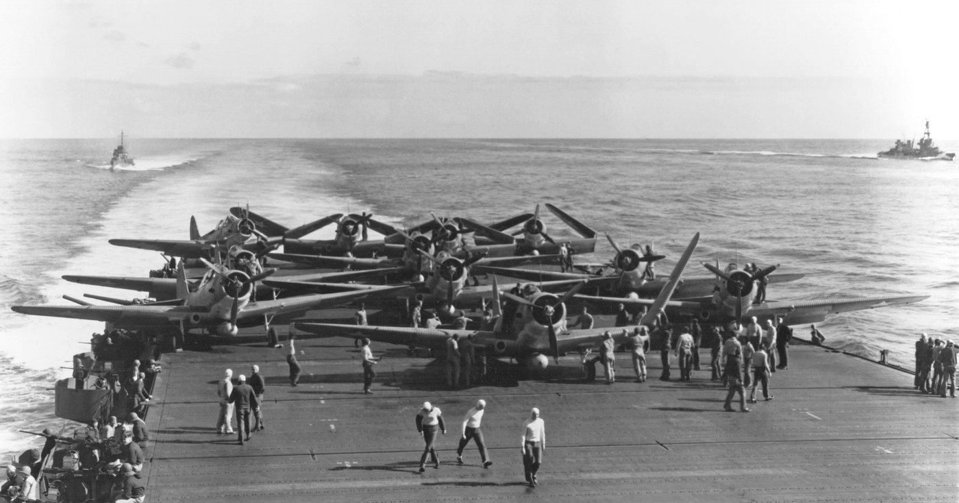 Black and white photograph of a group of torpedo bombers with folded wings parked at the stern of an aircraft carrier, with deck crew and escorting warships