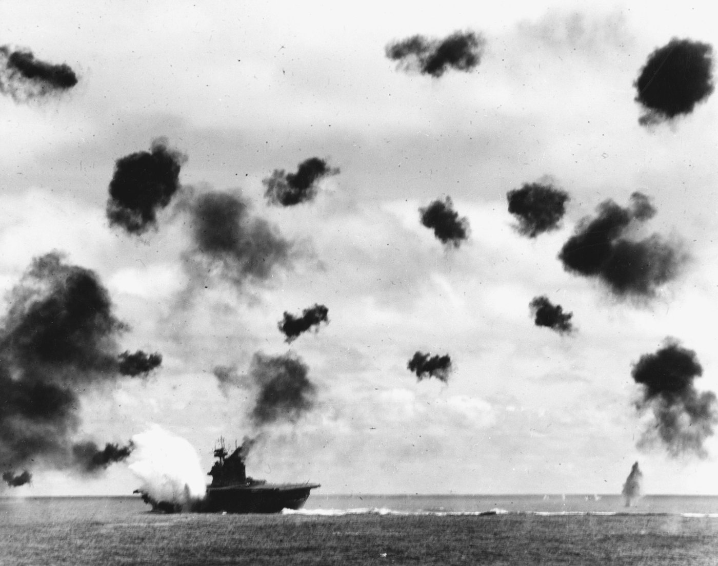 Black and white photograph of an aircraft carrier on the horizon with a white explosion at its side and dark flak bursts filling the sky