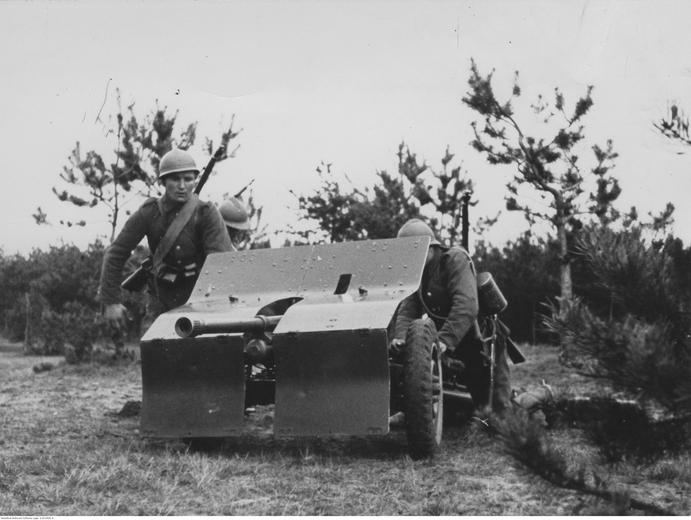 Black and white photograph of soldiers in helmets manning a small shielded anti-tank gun among young pines