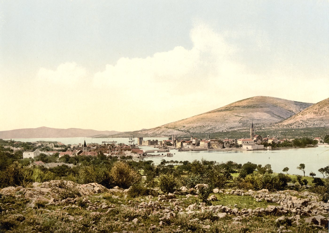 Color photochrom of a small town with a tall bell tower on a low island in a bay, below bare hills, seen across rocky scrubland
