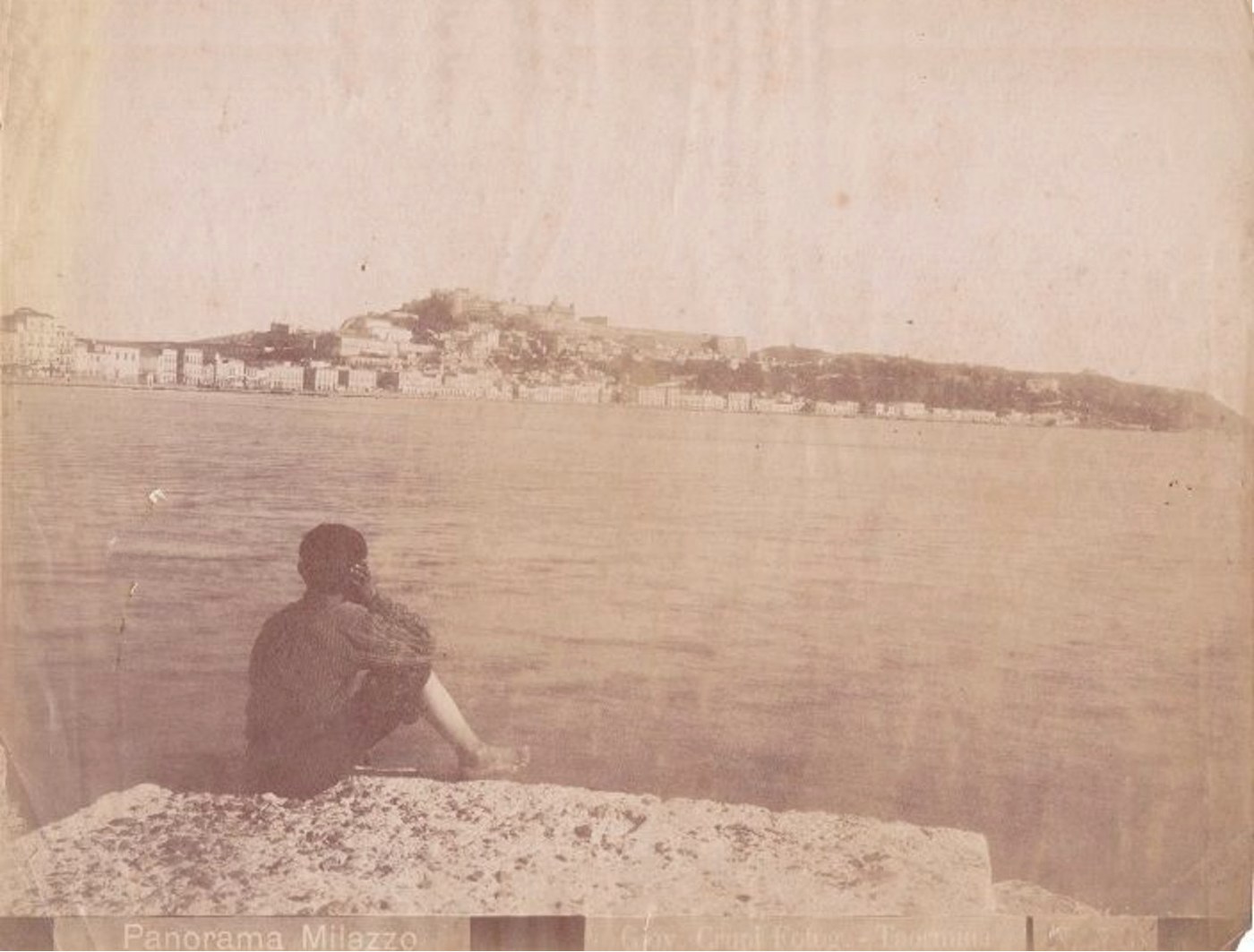 Wyblakła fotografia w sepii przedstawiająca spokojną zatokę z niskim miastem Milazzo na dalekim brzegu, zamkiem na wzgórzu nad nim i chłopcem siedzącym na kamieniu na pierwszym planie i patrzącym na wodę
