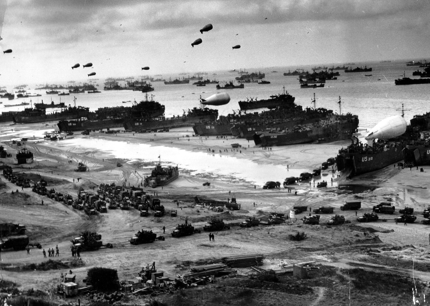 Landing ships and trucks crowding a Normandy beach with barrage balloons overhead