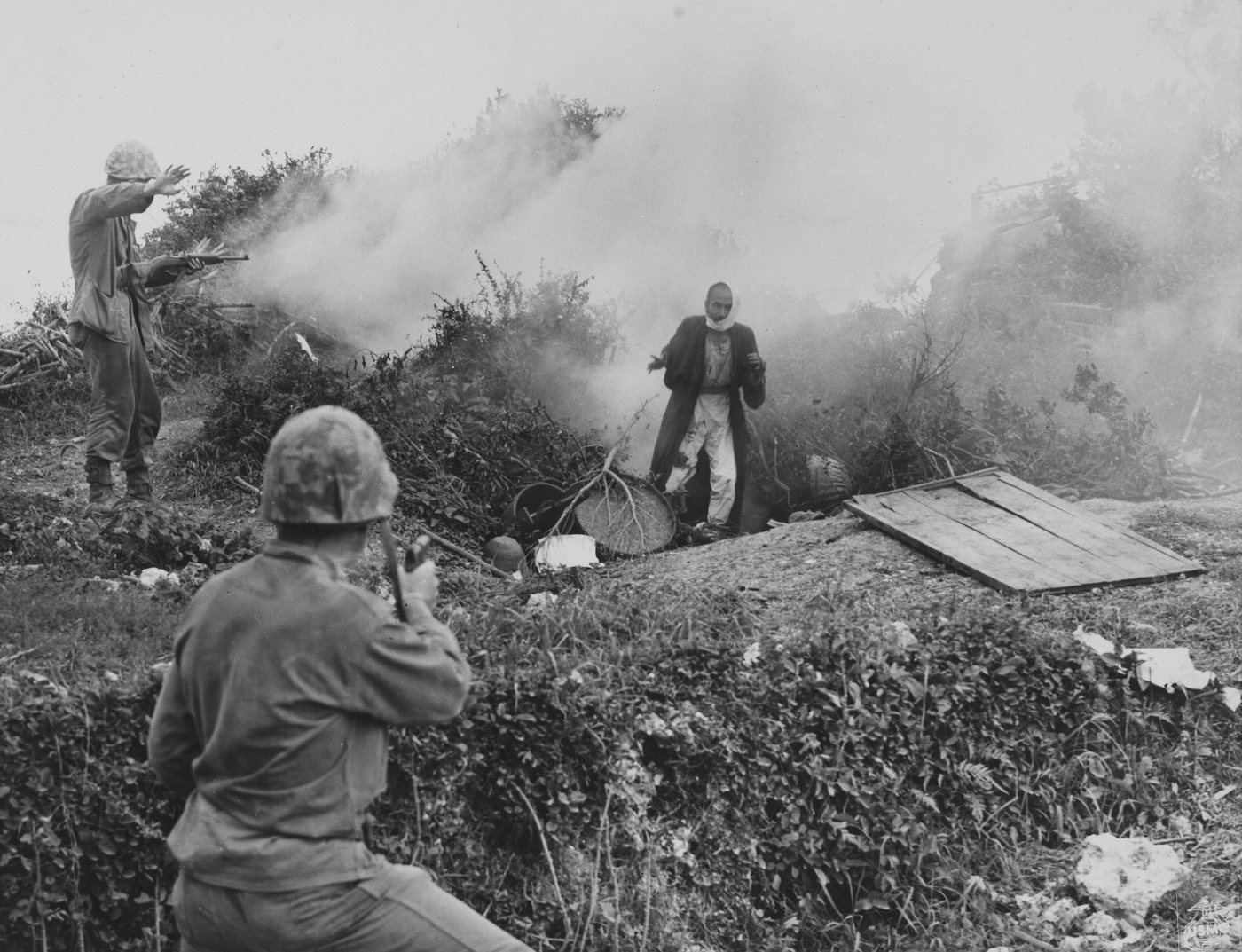 Marine with a raised arm on a hillside as a civilian walks out of a smoky cave