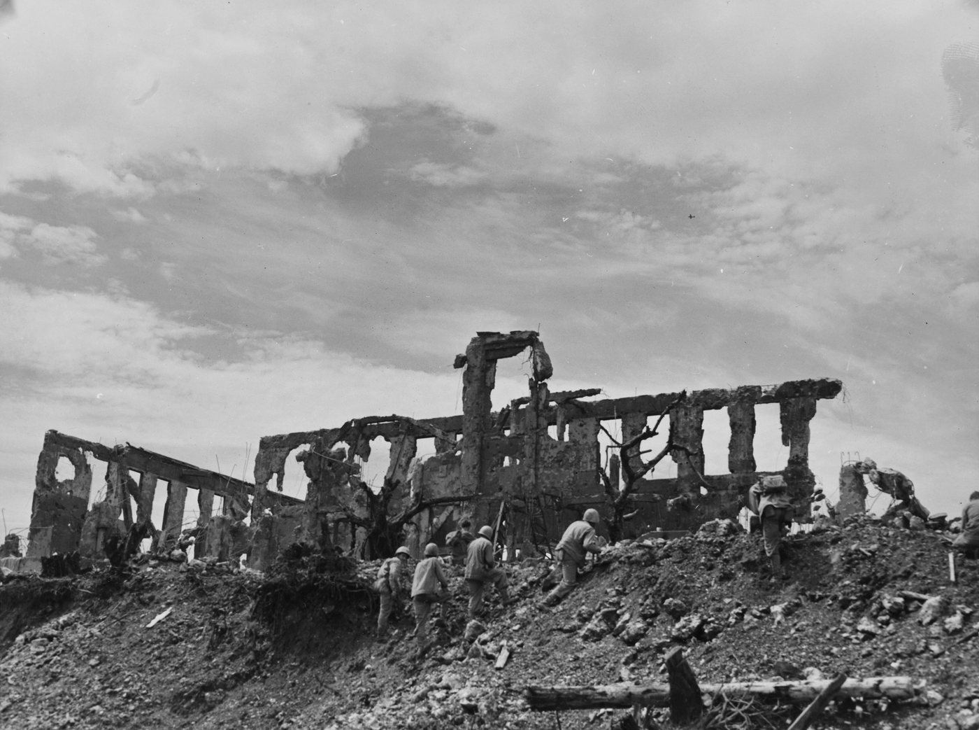 Marines advancing over rubble below the shattered walls of a ruined building