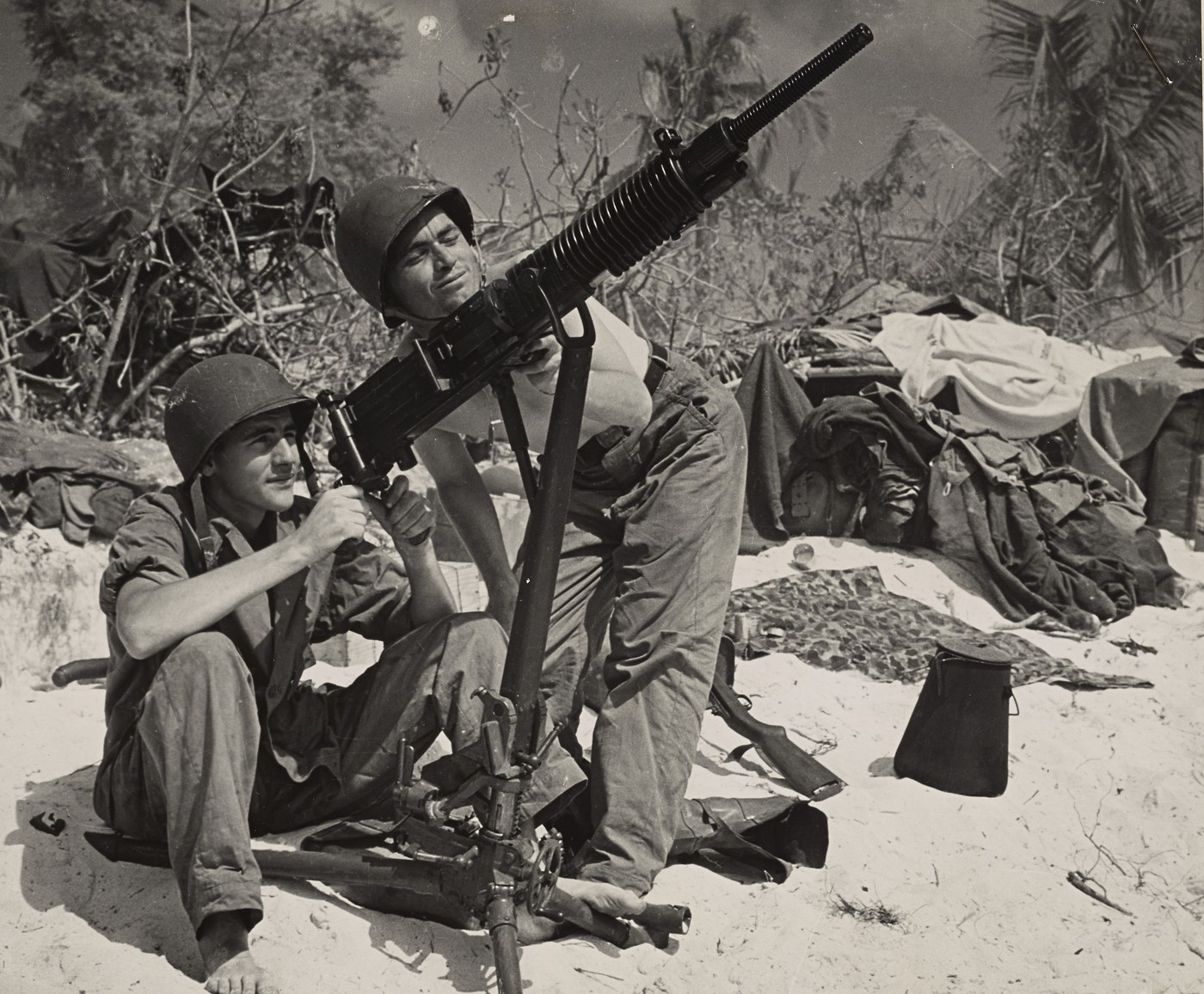 Black and white photograph of two young men in steel helmets on a sandy beach examining a captured machine gun on a tall tripod, one sitting barefoot gripping the gun, the other kneeling behind it and sighting along the barrel, with piled clothing, gear and shattered palm trees behind them