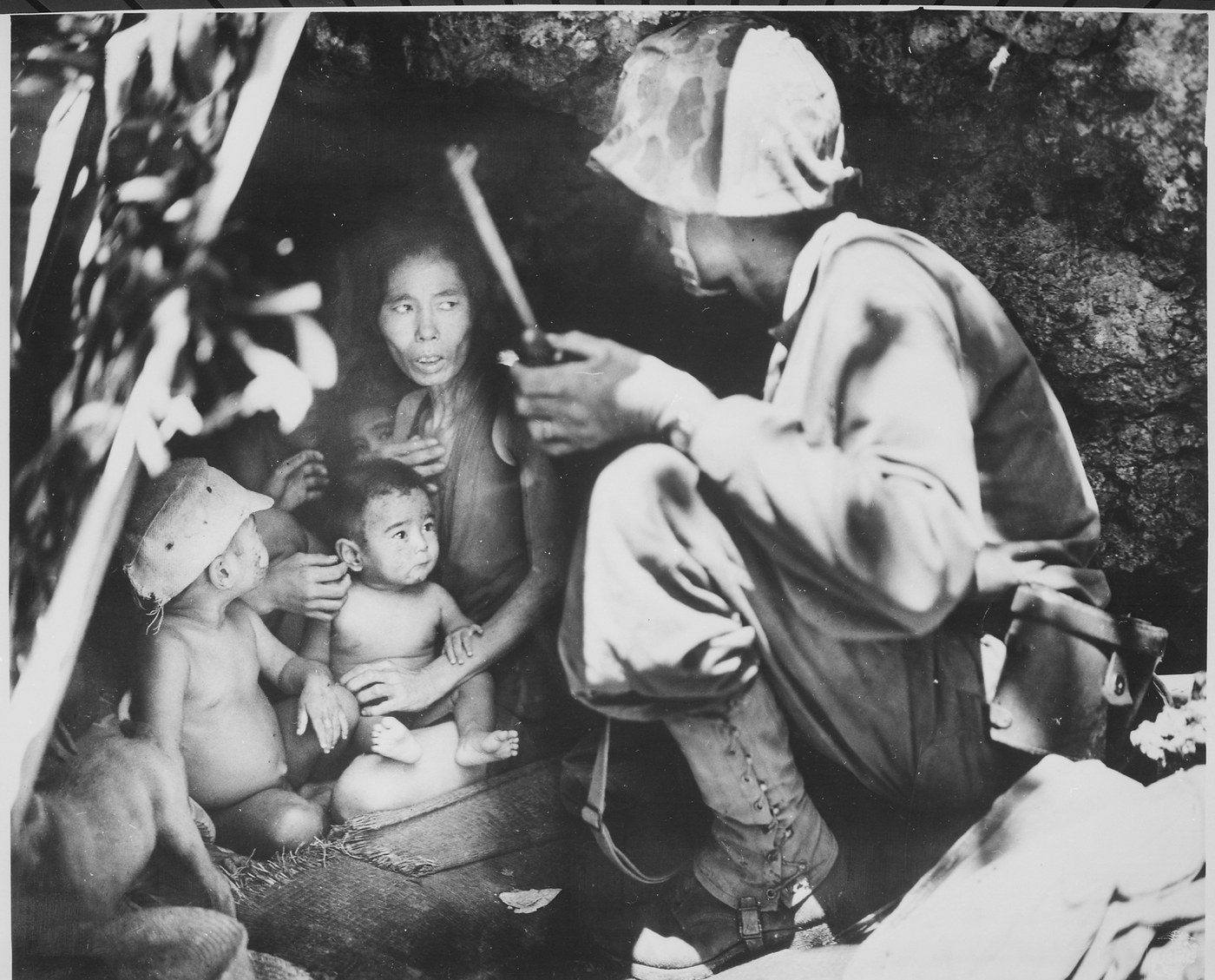 Black and white photograph of a Marine in a camouflage helmet cover crouching at the mouth of a rocky cave and speaking to a thin woman sitting inside with three small children, one of them a baby on her lap