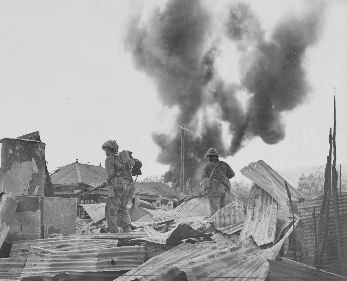 Black and white photograph of two US Marines with rifles and packs picking their way across a field of flattened corrugated iron sheets and wrecked buildings, a huge column of black smoke rising behind a utility pole