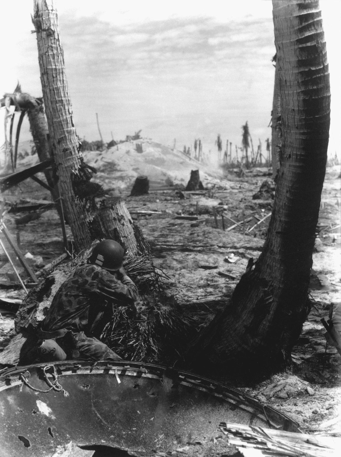 Photograph of a Marine crouching behind a broken palm trunk in a devastated landscape