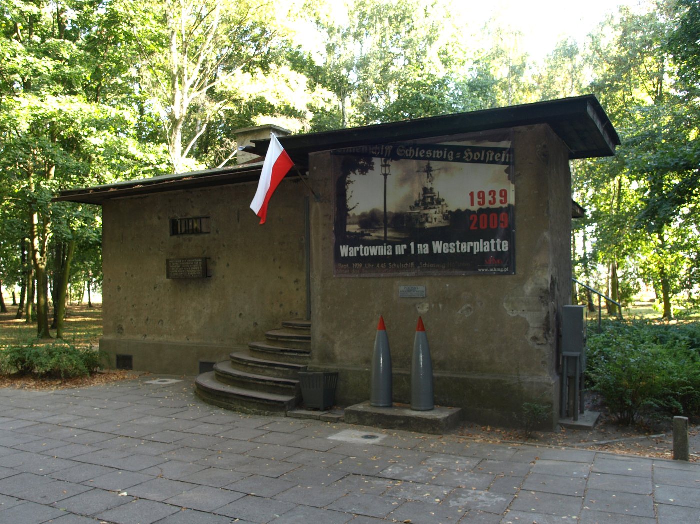 Small concrete guardhouse among trees with a Polish flag and two large shells by the steps