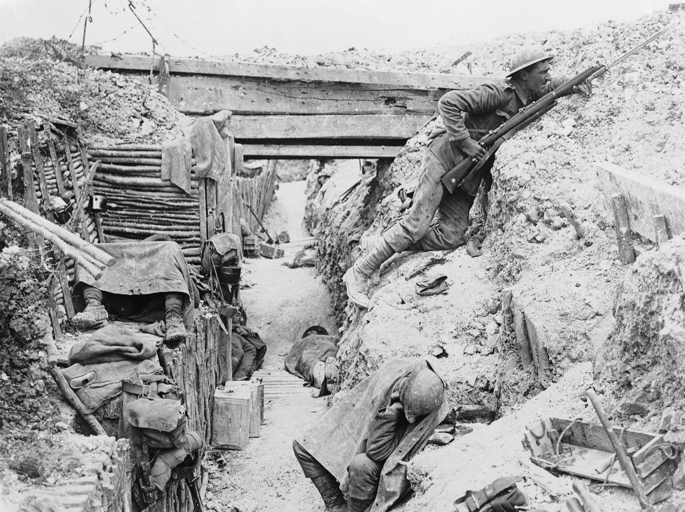 Black and white photograph of a captured German trench: one soldier keeps watch over the back wall of the trench with a rifle while others rest below.