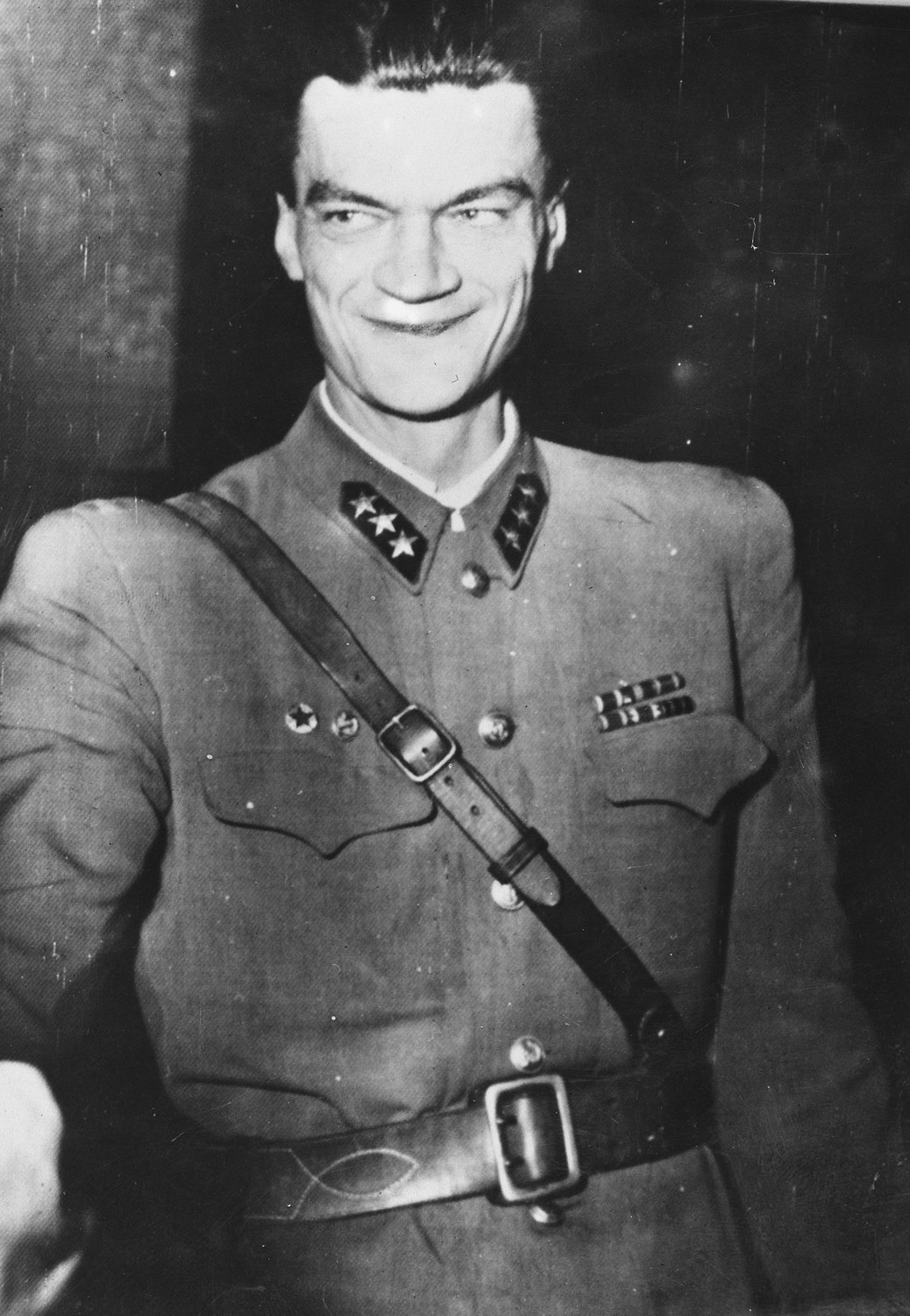 Black and white portrait of a smiling officer with a high forehead and swept back hair, wearing a uniform tunic with a cross strap, collar insignia with stars and ribbon bars, shown on a creased press print